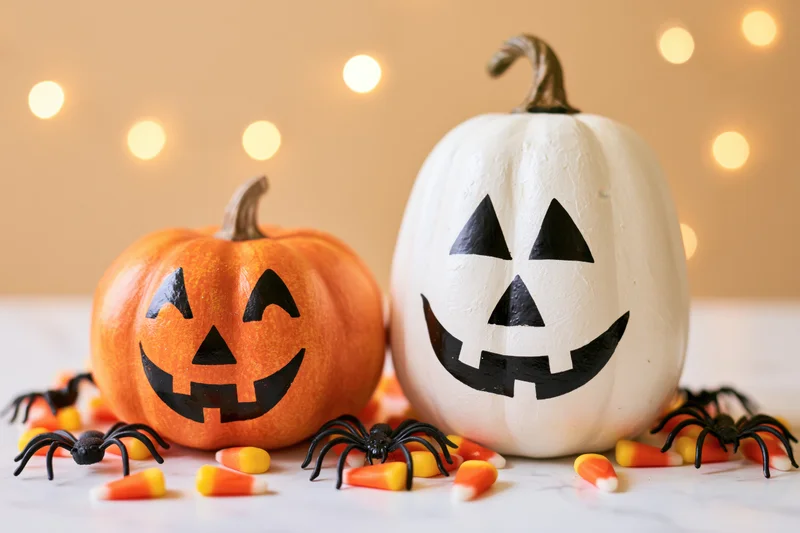 Two painted jack-o-lantern pumpkins with smiling faces on a table surrounded by candy corn and plastic spiders, warm lights in background