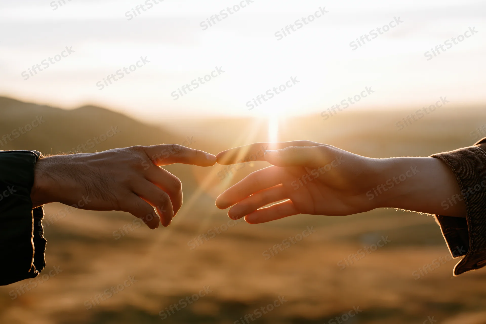 Two hands reaching toward each other with fingertips almost touching against a sunset landscape