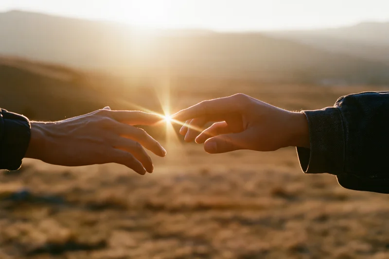 Two hands reaching toward each other with fingertips almost touching at sunset in a mountainous landscape