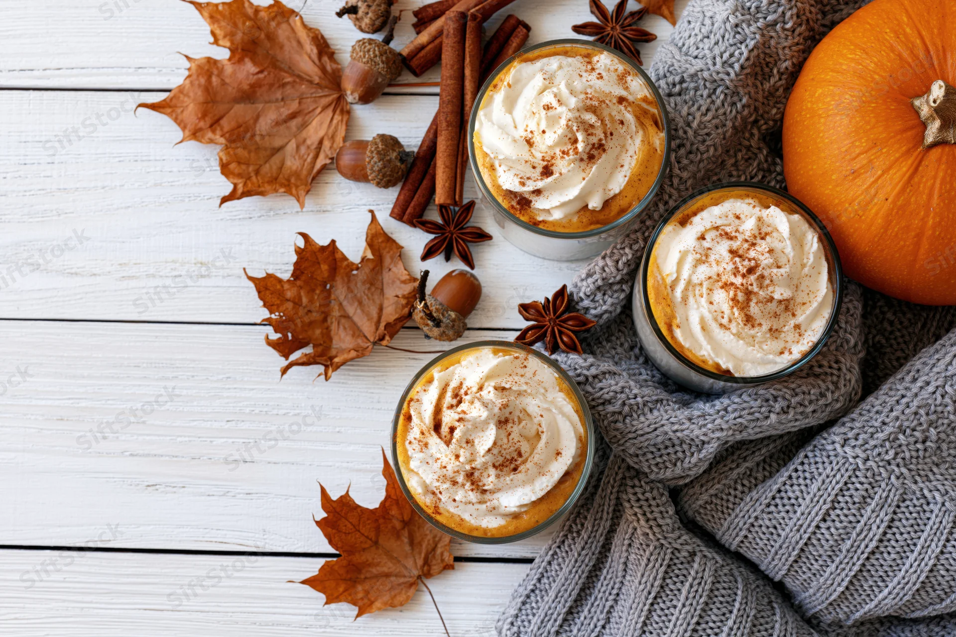 Three pumpkin spice lattes with whipped cream and cinnamon on a white wooden table surrounded by autumn leaves, acorns, cinnamon sticks, star anise, a knitted sweater, and a pumpkin