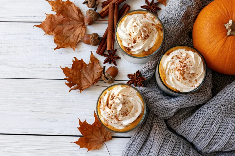 Three pumpkin spice lattes with whipped cream and cinnamon on a white wooden table surrounded by autumn leaves, acorns, cinnamon sticks, star anise, a knitted sweater, and a pumpkin