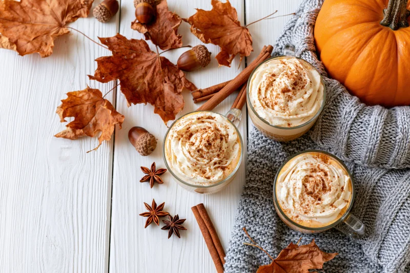 Three pumpkin spice lattes with whipped cream and cinnamon on a white wooden table surrounded by autumn leaves, acorns, star anise, cinnamon sticks, and a gray knitted sweater