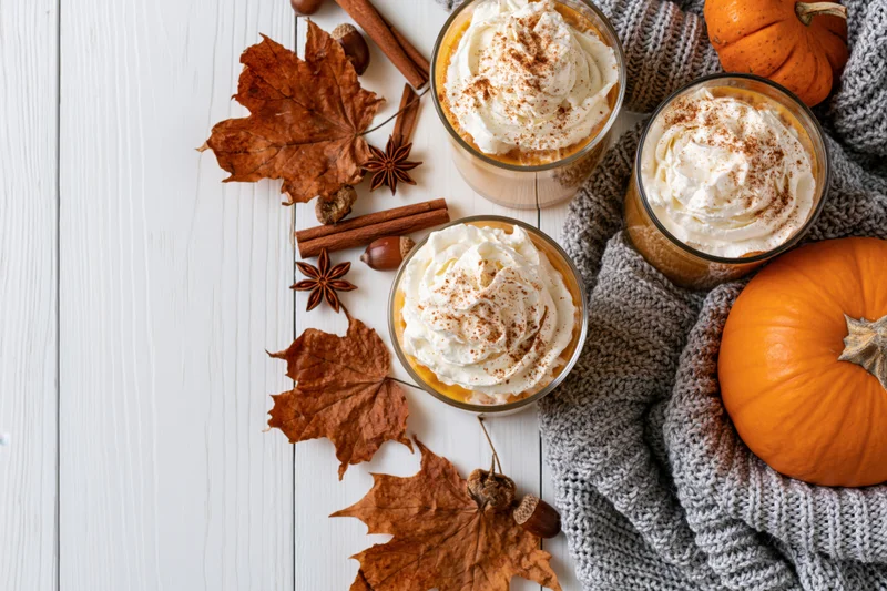 Three pumpkin spice lattes with whipped cream and cinnamon on a white wooden table beside autumn leaves, pumpkins, and a gray knitted sweater
