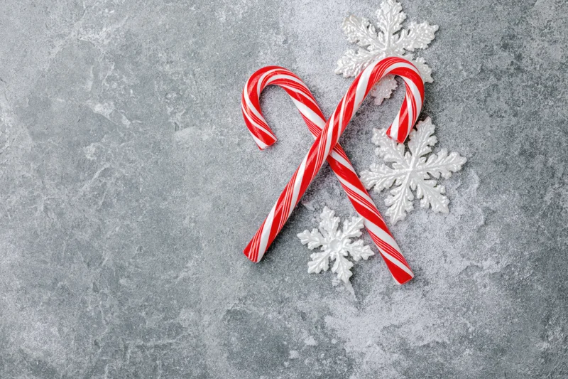 Two red and white striped candy canes crossed on a gray textured surface with white snowflake ornaments