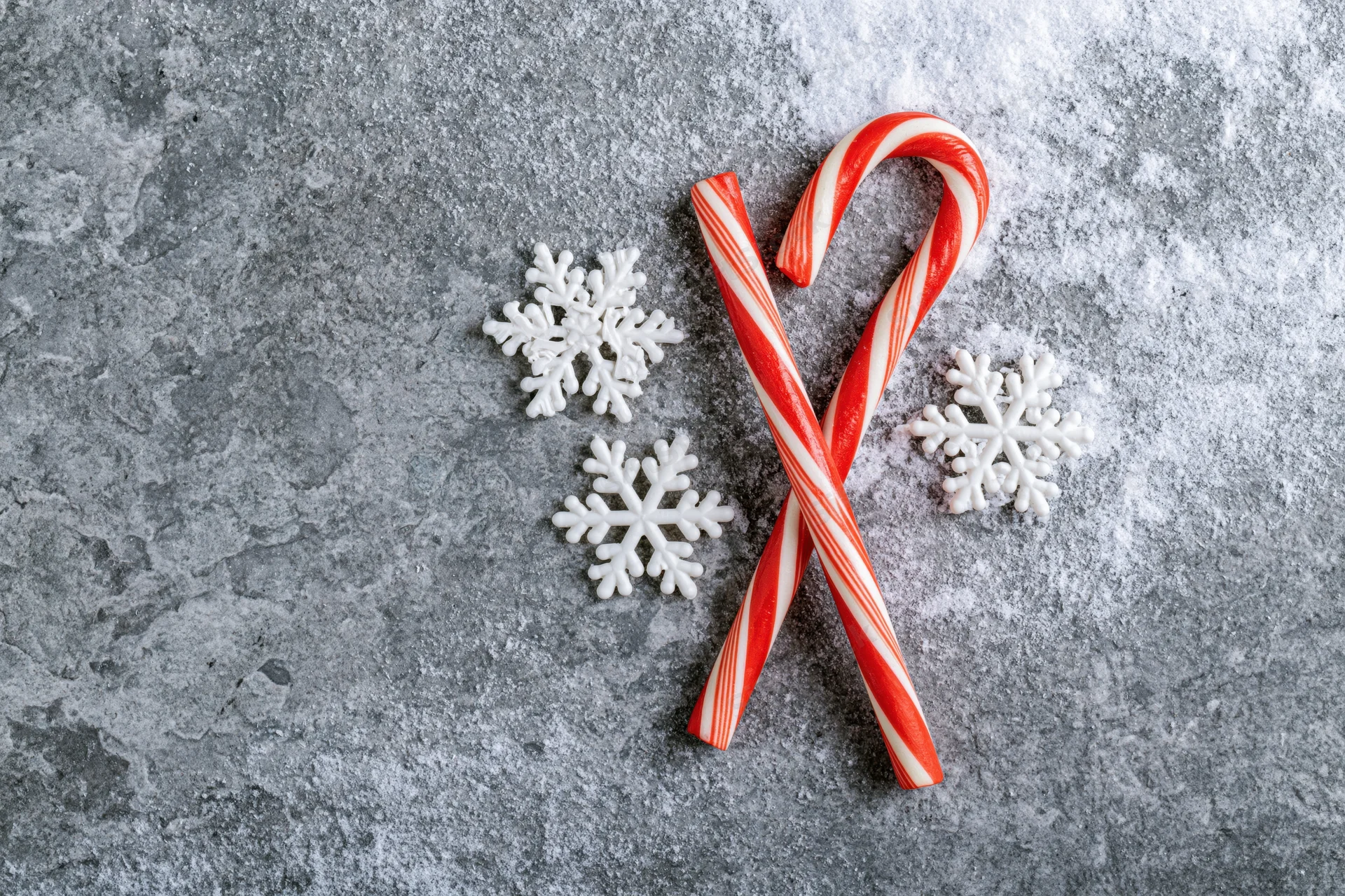 Two red and white striped candy canes crossed on a gray textured surface with white snowflake ornaments around them