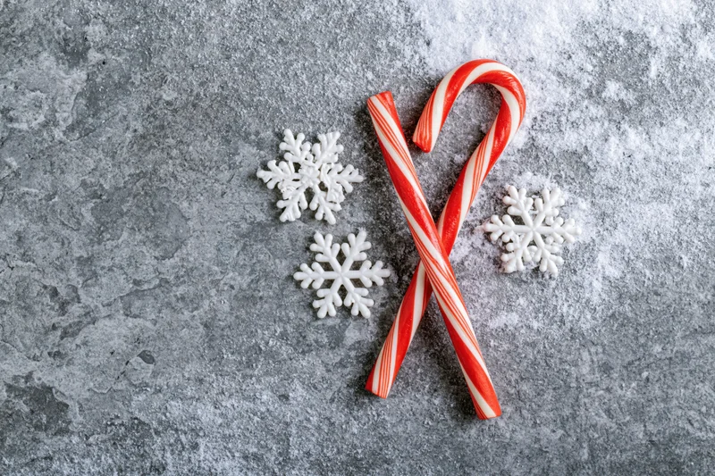 Two red and white striped candy canes crossed on a gray textured surface with white snowflake ornaments around them