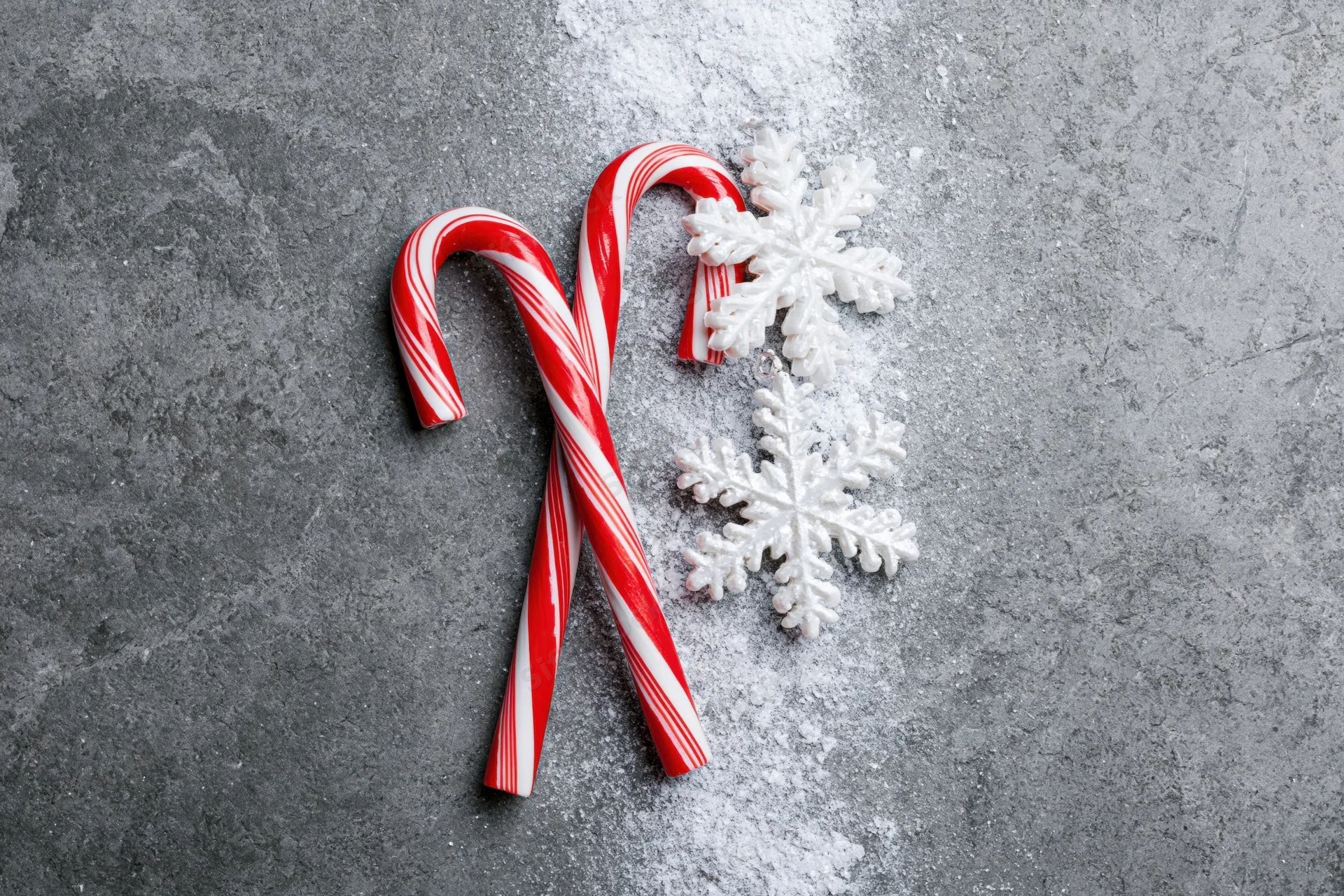 Two red and white striped candy canes crossed on a gray textured surface with white snowflakes and scattered powder