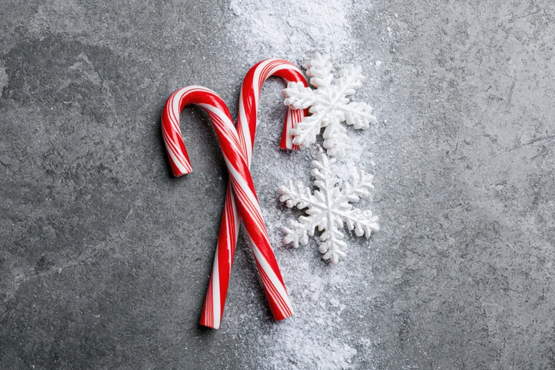 Two red and white striped candy canes crossed on a gray textured surface with white snowflakes and scattered powder
