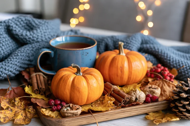 Two small orange pumpkins on a wooden tray surrounded by dried leaves, berries, cinnamon sticks, pinecones, and a blue mug of tea, set on a knitted blanket with warm string lights in the background