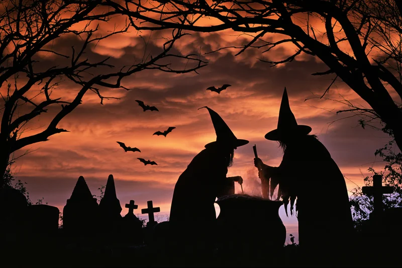 Two witches in pointed hats stirring a cauldron in a graveyard at twilight