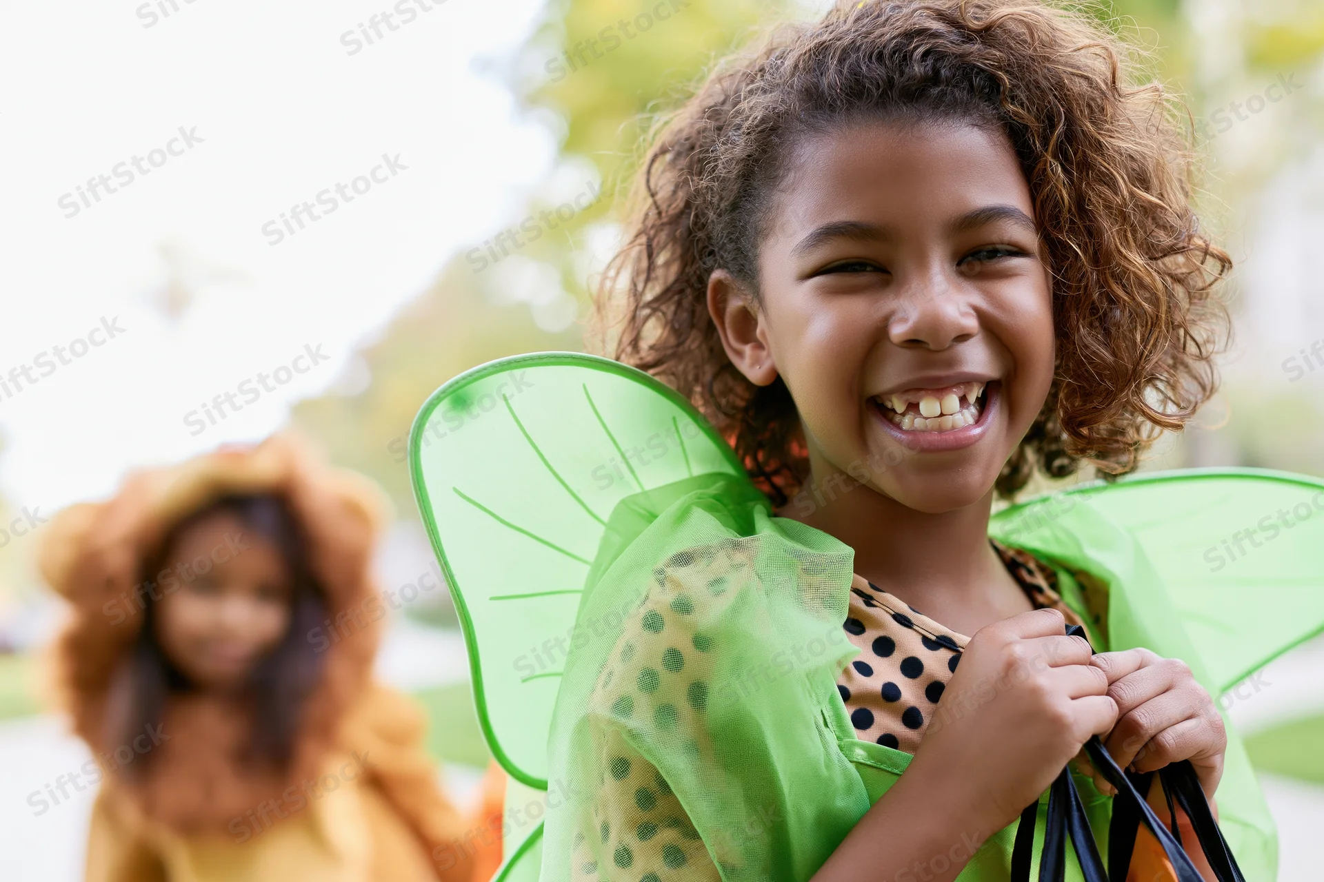 Young black girl in green fairy costume smiling while holding trick-or-treat bag outdoors