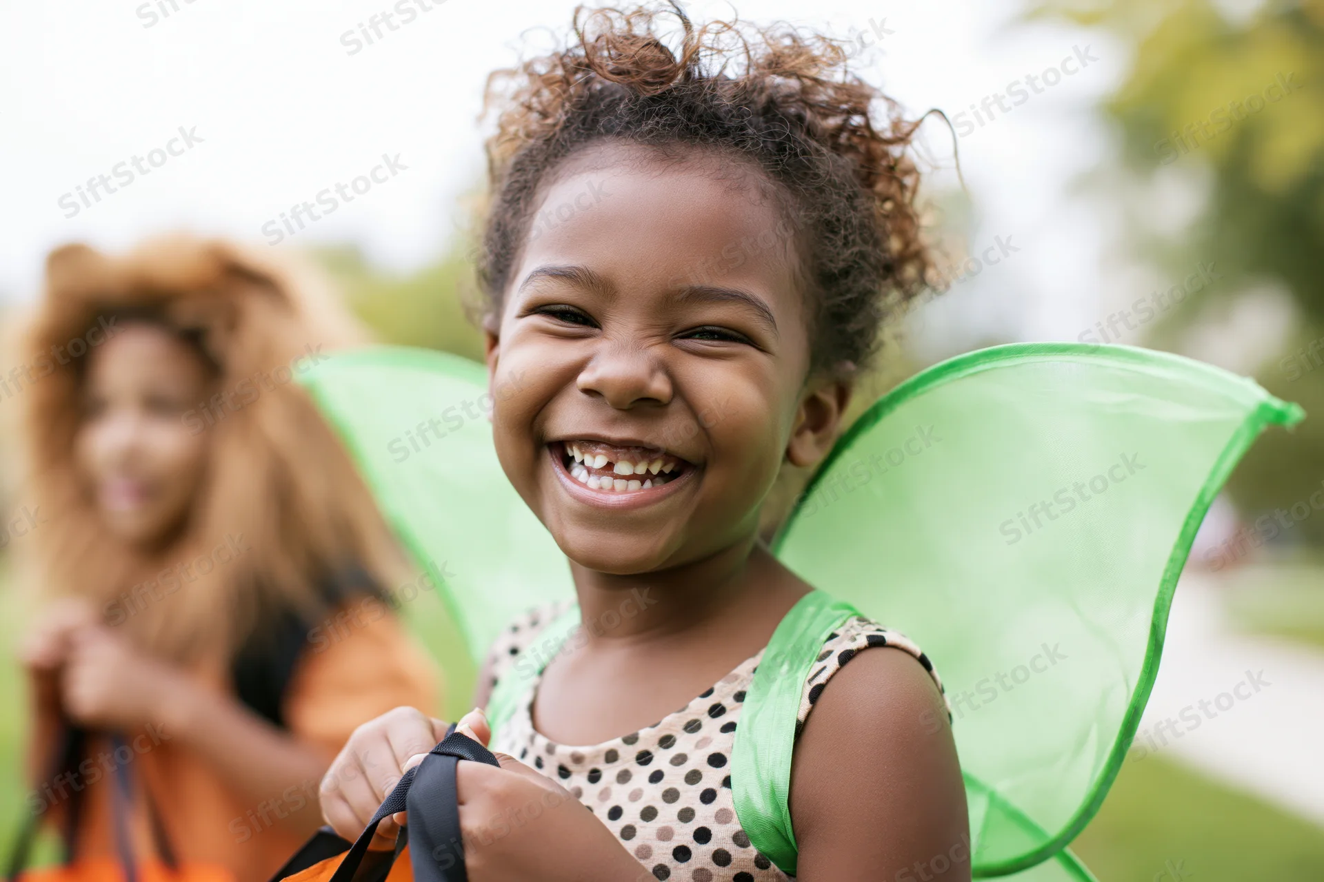 Young black girl smiling in green fairy wings costume outdoors with another child in lion costume behind her