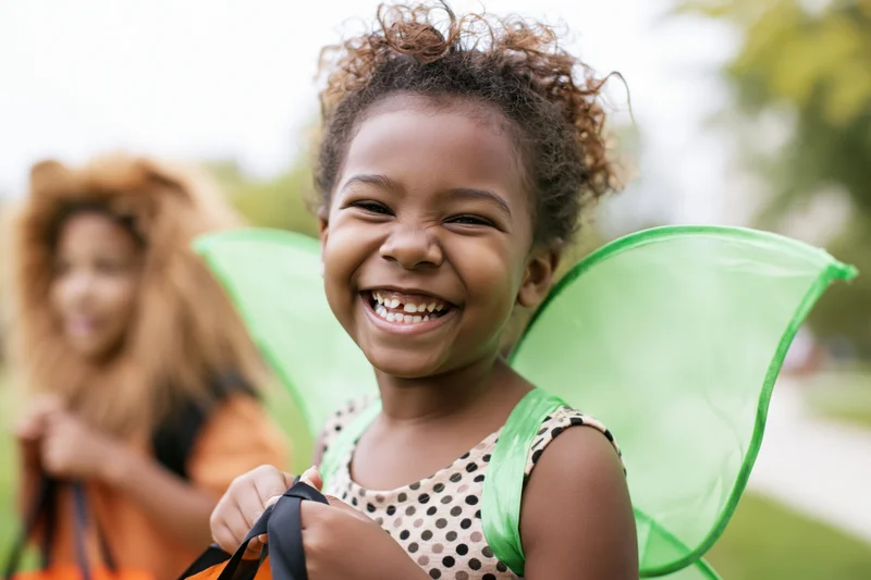 Young black girl smiling in green fairy wings costume outdoors with another child in lion costume behind her