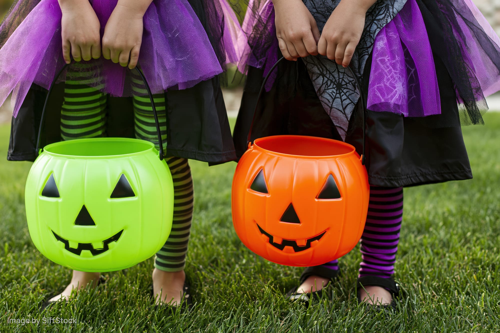 Two young girls in witch costumes holding plastic pumpkin buckets on grass during halloween