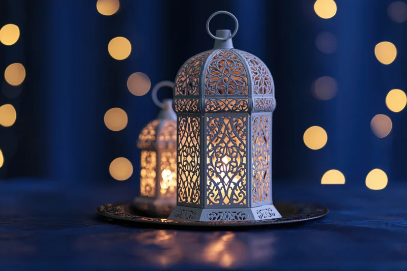 White ornate moroccan lantern with lit candle on tray against dark blue background with golden bokeh lights