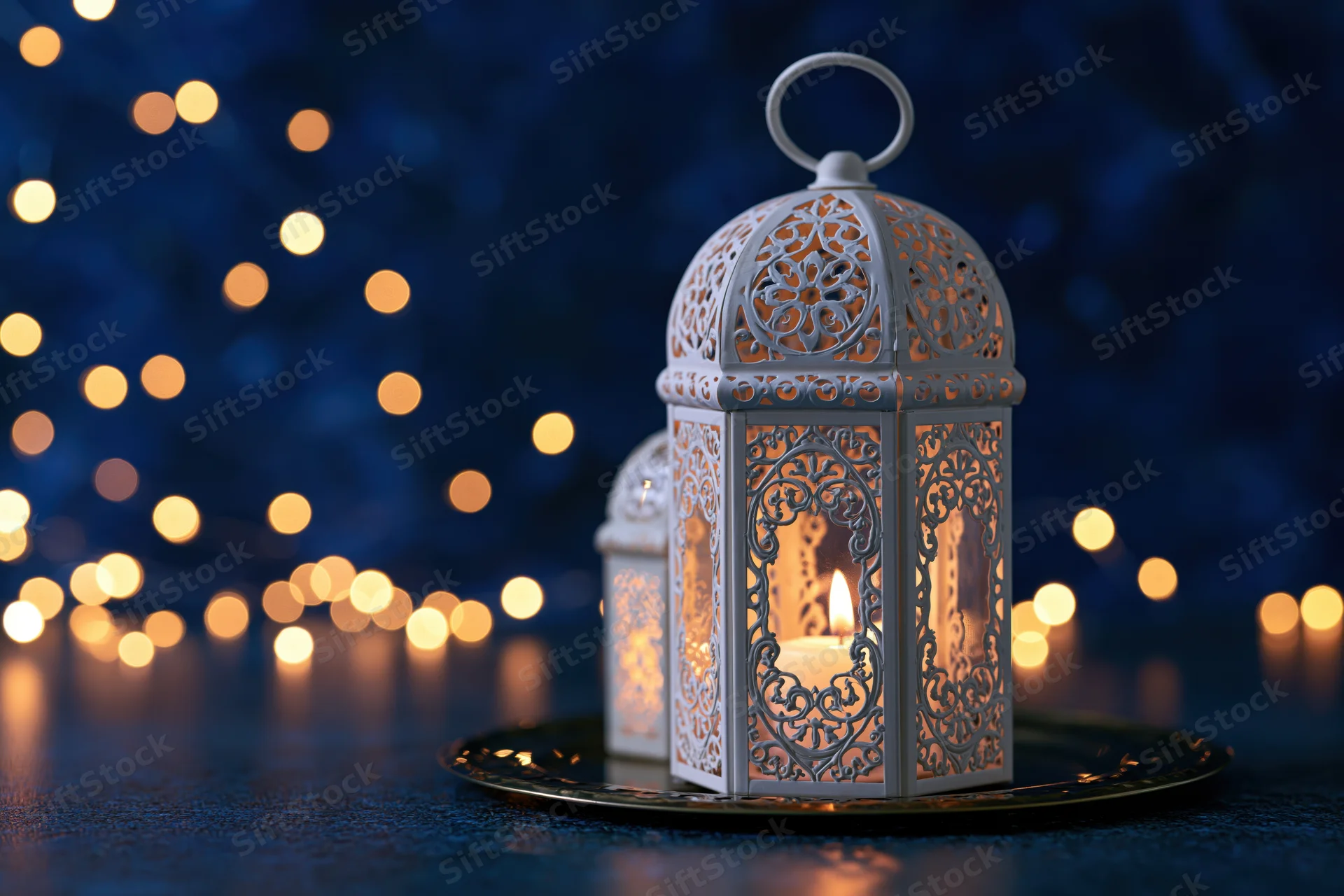 White ornate moroccan lantern with lit candle on dark surface against bokeh lights at night