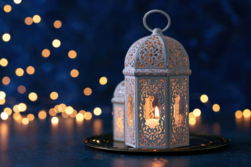 White ornate moroccan lantern with lit candle on dark surface against bokeh lights at night