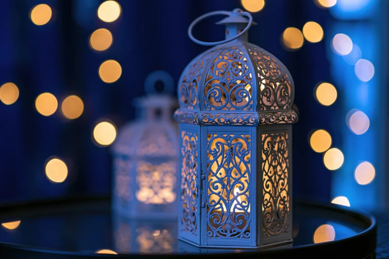 White ornate moroccan lantern with lit candle on dark reflective surface against blue bokeh lights