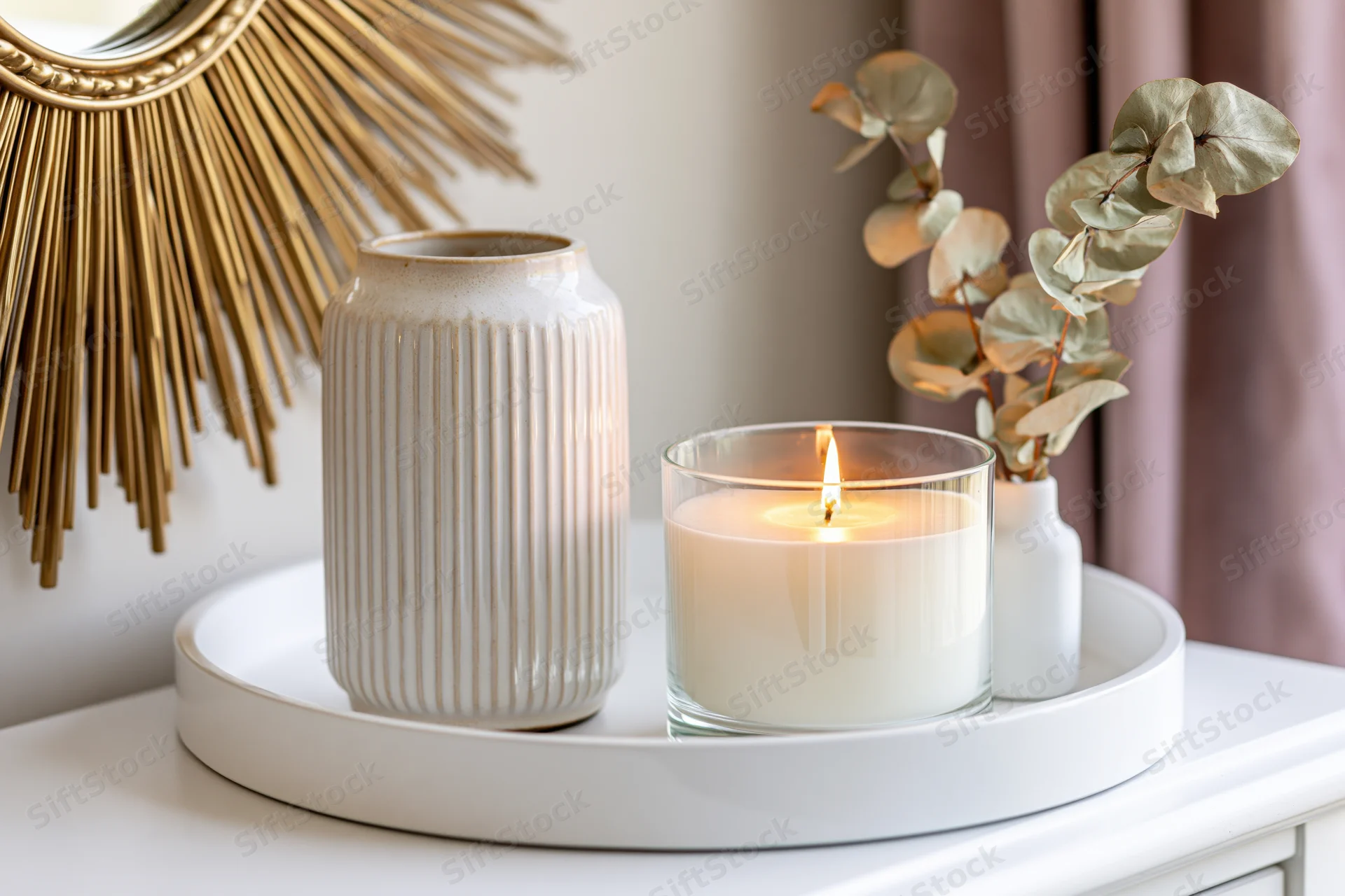 Lit candle and ribbed ceramic vase on white tray beside sunburst mirror on sideboard