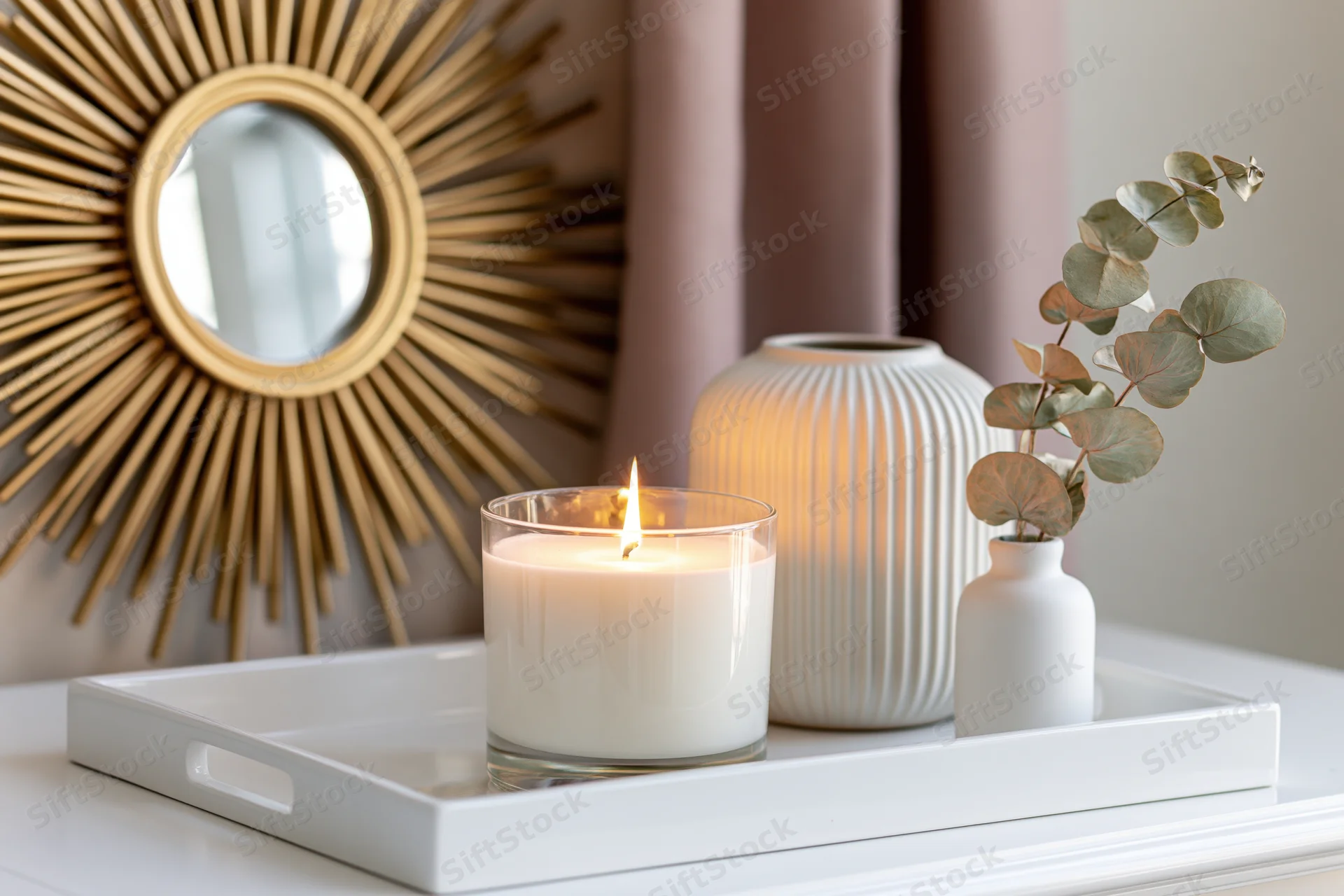 Lit candle and ceramic vase on white tray with eucalyptus branch and sunburst mirror in background