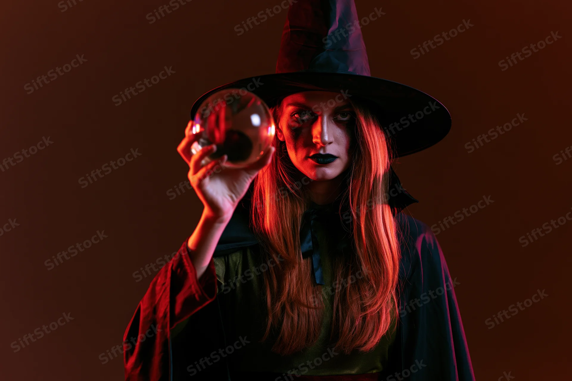 Woman in witch costume holding crystal ball under red lighting in studio