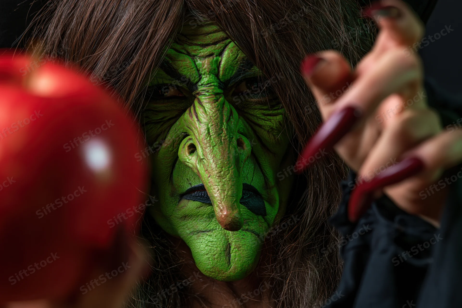 Closeup of a woman in a green-skinned witch costume holding a red apple with long red nails visible