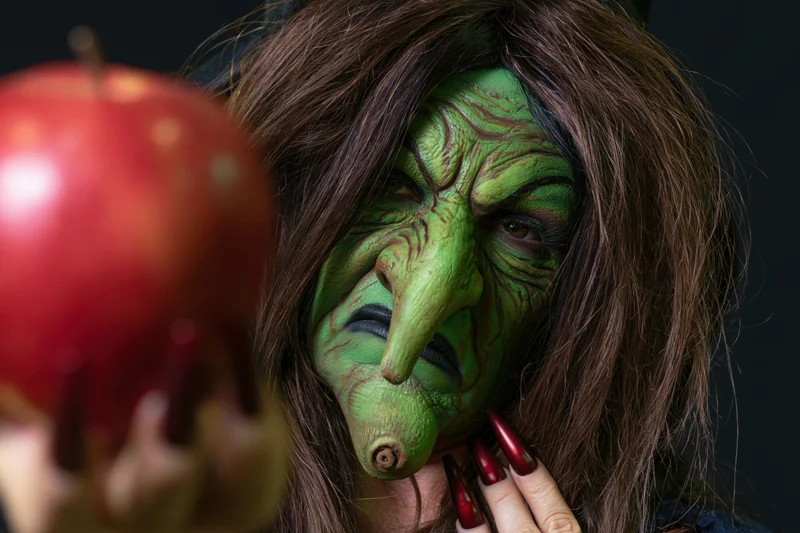 Woman in green witch makeup holding a red apple, close-up against black background