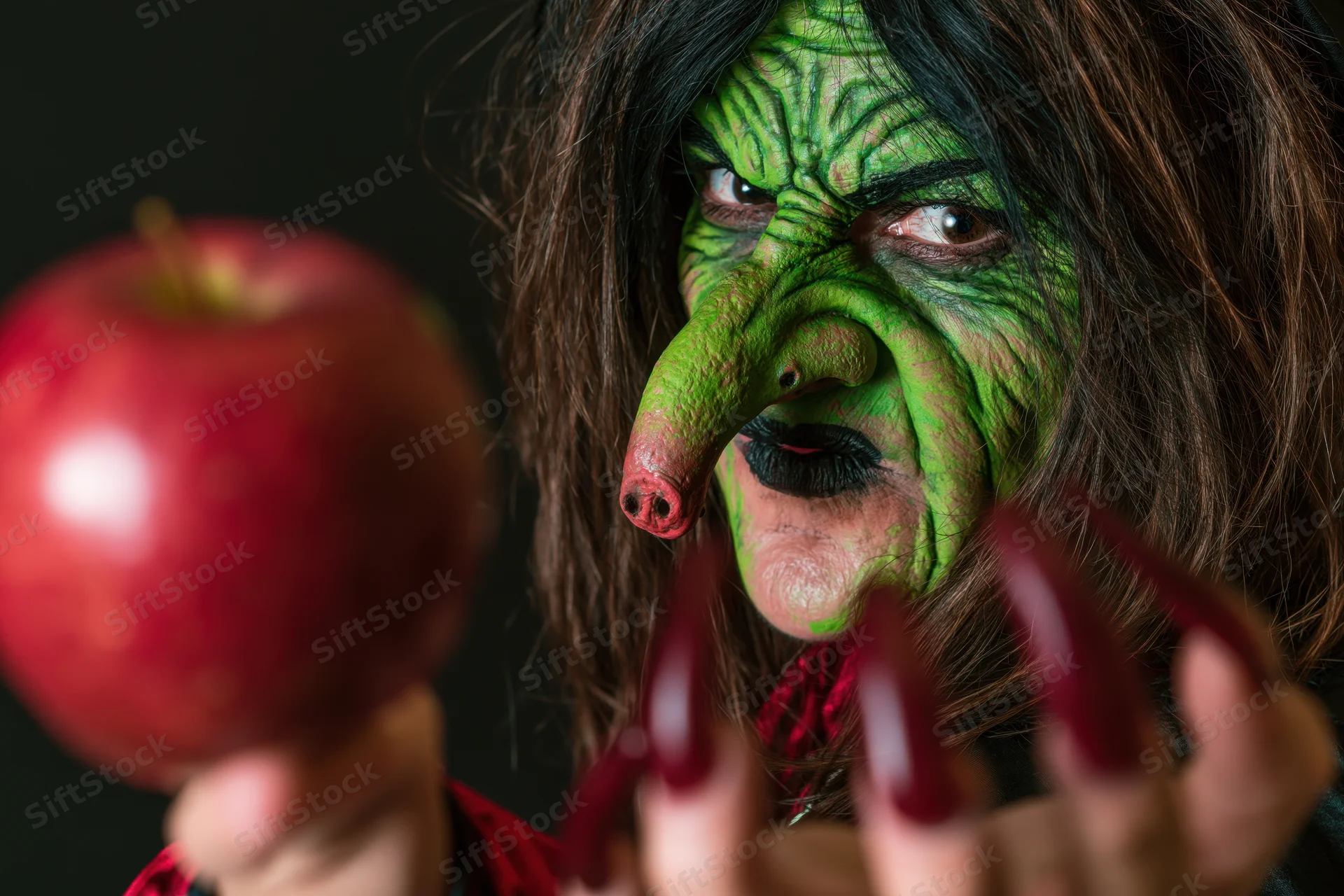 Woman in green witch makeup holding red apple with long red nails extended toward camera