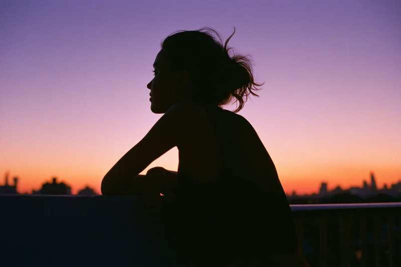 Woman in silhouette leaning on rooftop railing gazing at cityscape at sunset