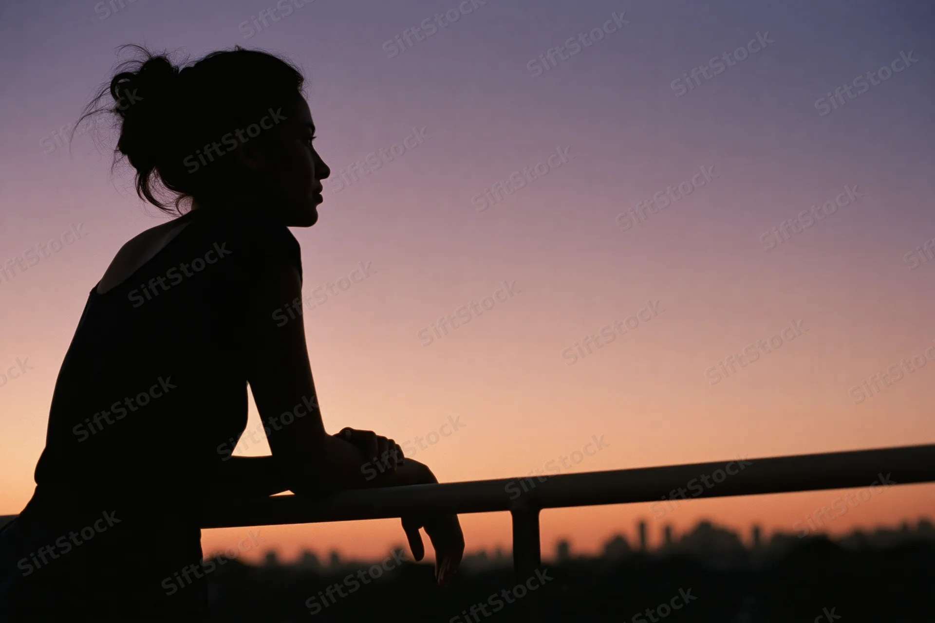 Silhouette of a woman leaning on a rooftop railing gazing into the distance at sunset