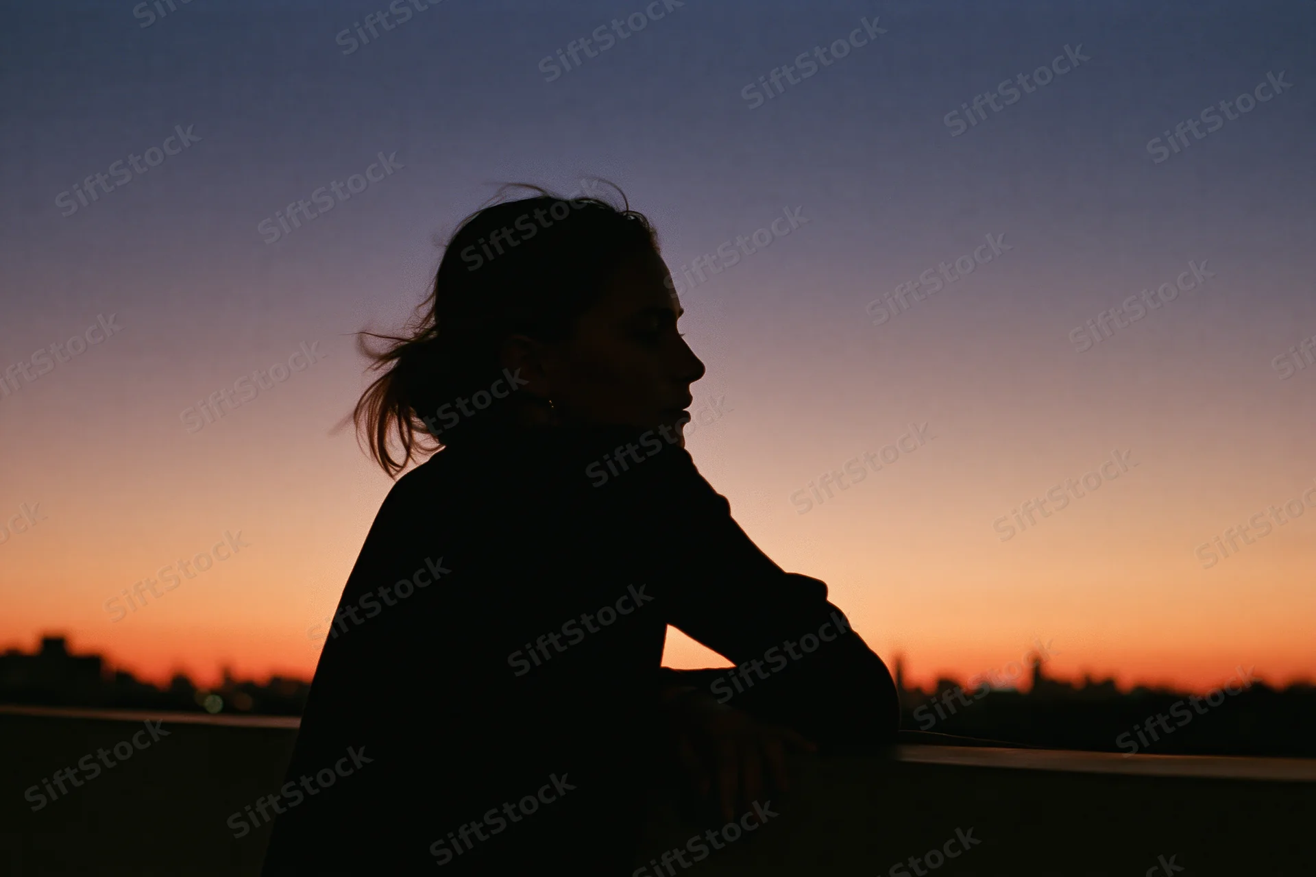 Woman in silhouette leaning on rooftop railing gazing into the distance at sunset