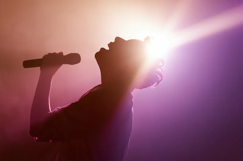 Woman singing into handheld microphone with head tilted back under stage spotlight