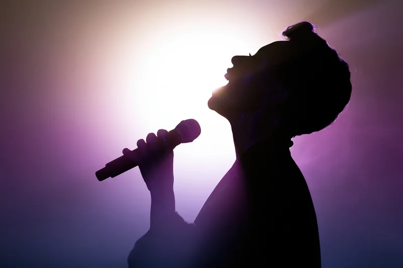 Silhouette of a woman singing into a microphone with head tilted back on stage under spotlight