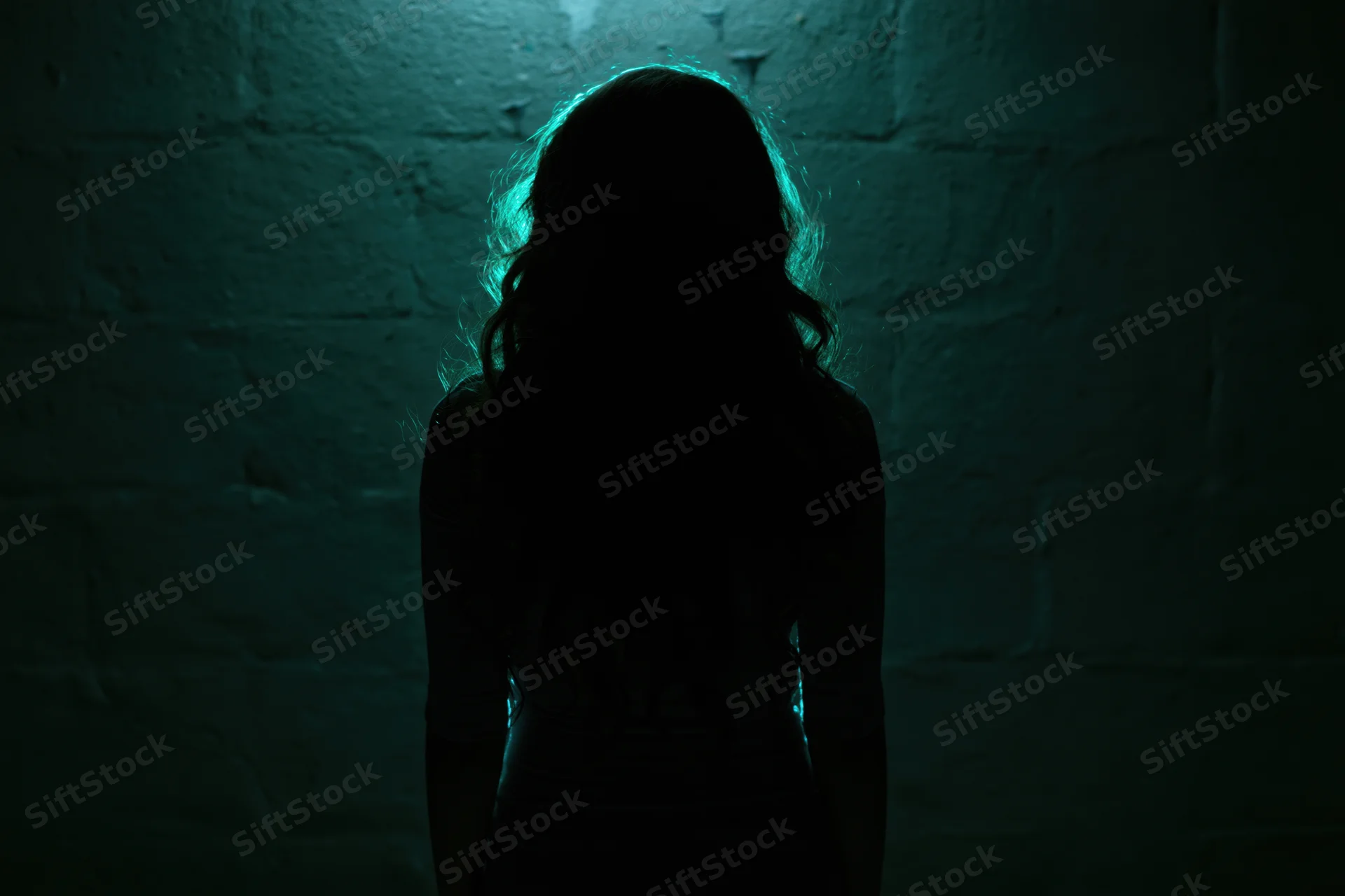 Woman in silhouette standing still facing away from camera against a brick wall in dim teal light