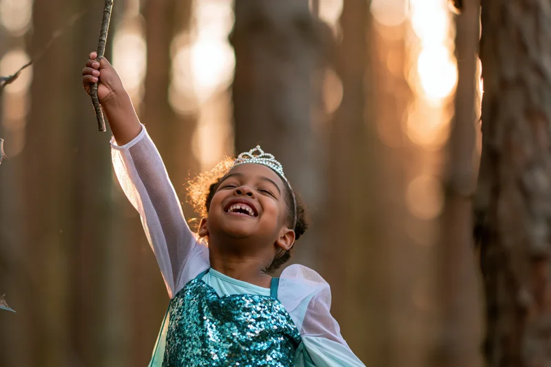 Young black girl laughing while holding a stick aloft in a sunlit forest, wearing a sparkly teal princess costume with tiara