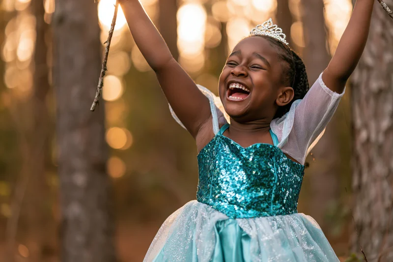 Young black girl laughing with arms raised holding a stick in a sunlit forest wearing a blue sequined princess dress and crown