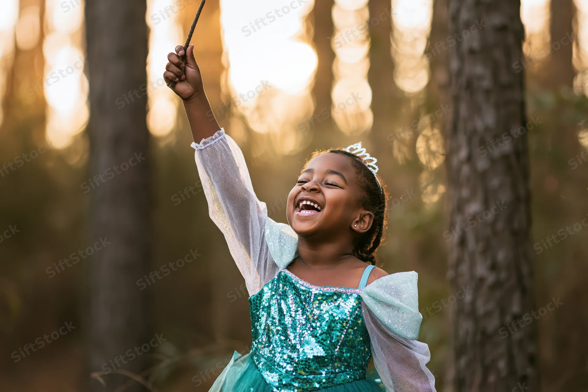 Young black girl laughing and raising a stick in a sunlit forest while wearing a teal sequin dress and tiara