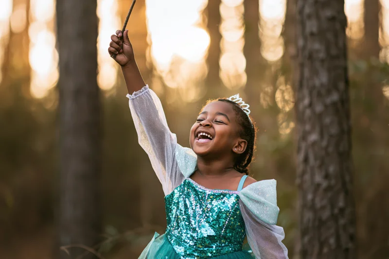 Young black girl laughing and raising a stick in a sunlit forest while wearing a teal sequin dress and tiara