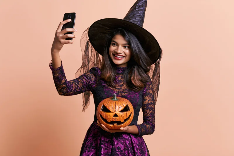 Young southasian woman in witch costume taking selfie with pumpkin against peach background