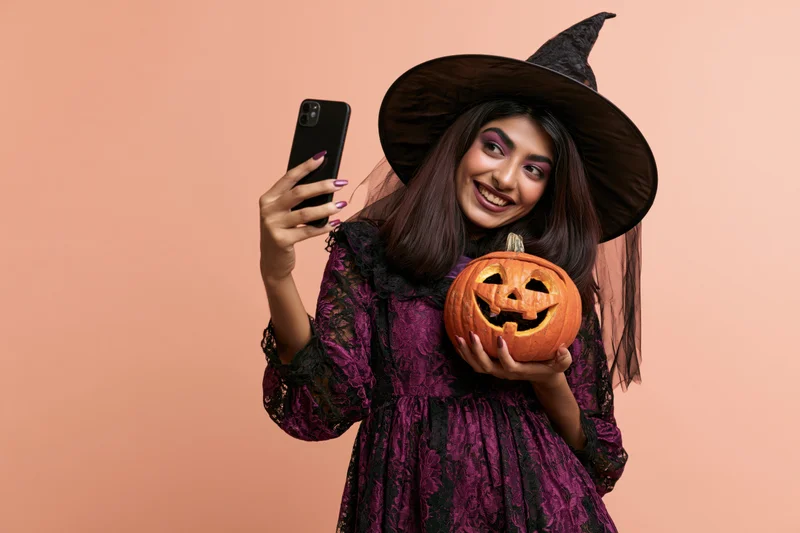 Young southasian woman in witch costume taking selfie with carved pumpkin in studio