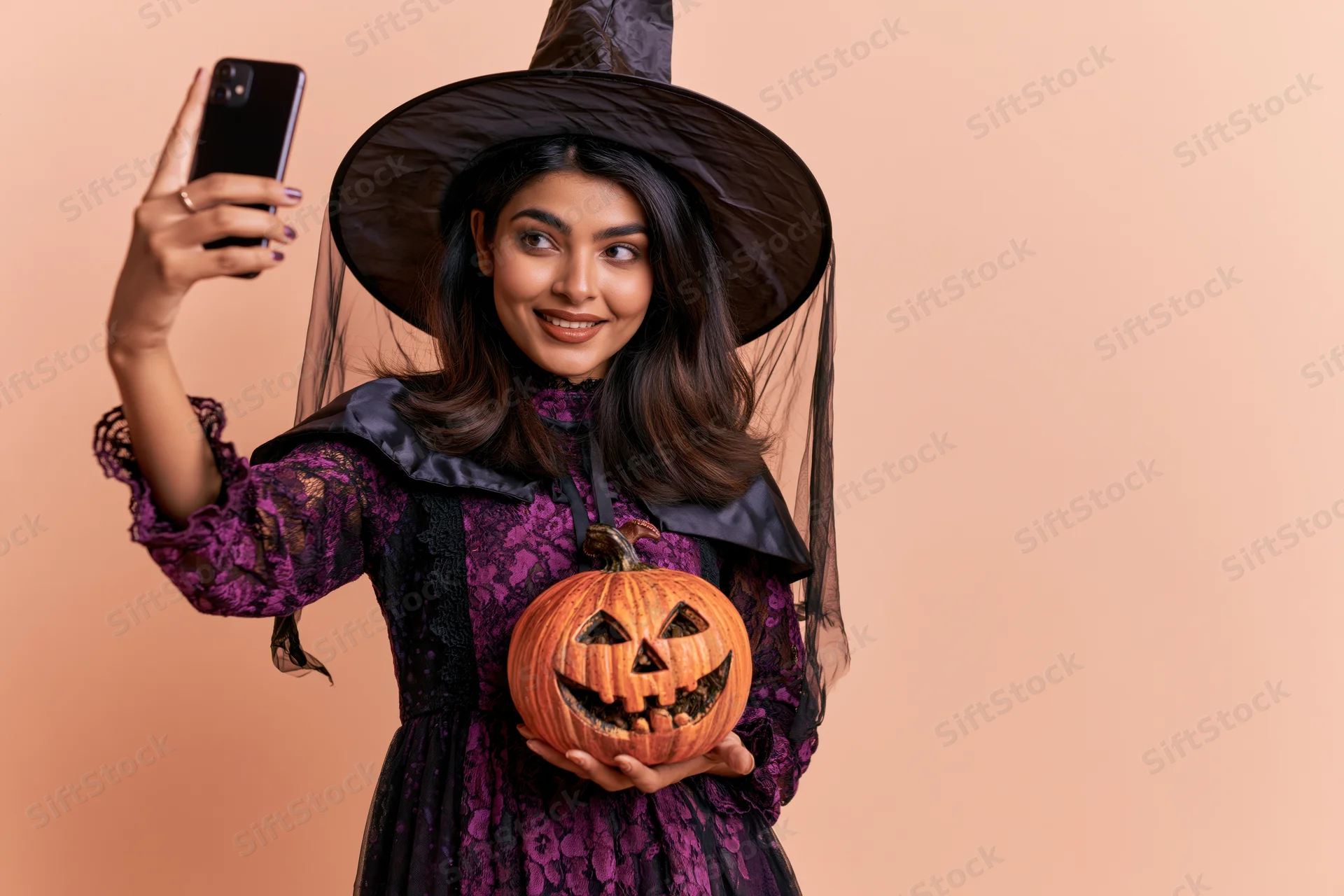 Young southasian woman in witch costume taking selfie with carved pumpkin against peach background