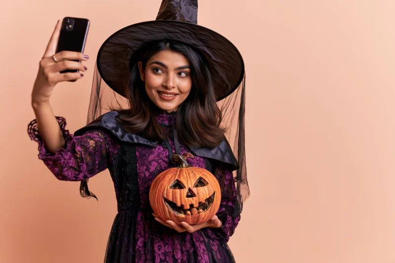 Young southasian woman in witch costume taking selfie with carved pumpkin against peach background
