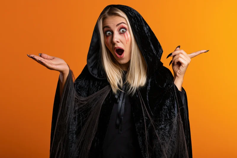 Young blonde woman in witch costume with open mouth and surprised expression against orange background