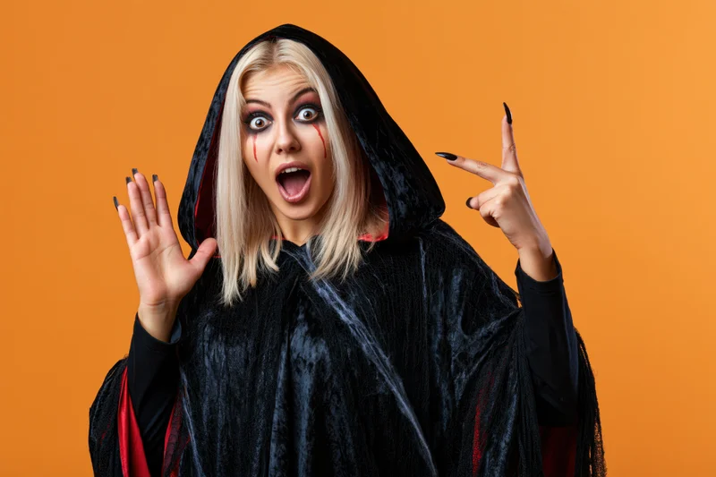 Blonde woman in black hooded cloak with red lining, making surprised expression and hand gestures against orange background