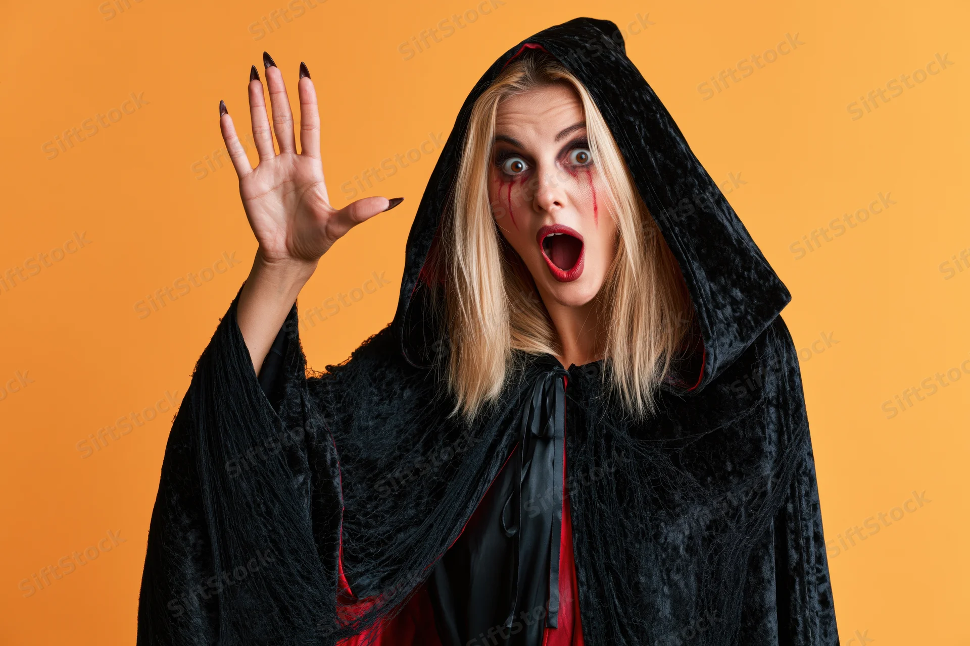 Blonde woman in witch costume with open mouth and raised hand against orange background