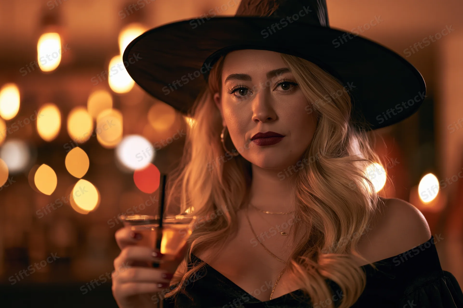 Blonde woman in black witch costume holding cocktail at halloween party indoors