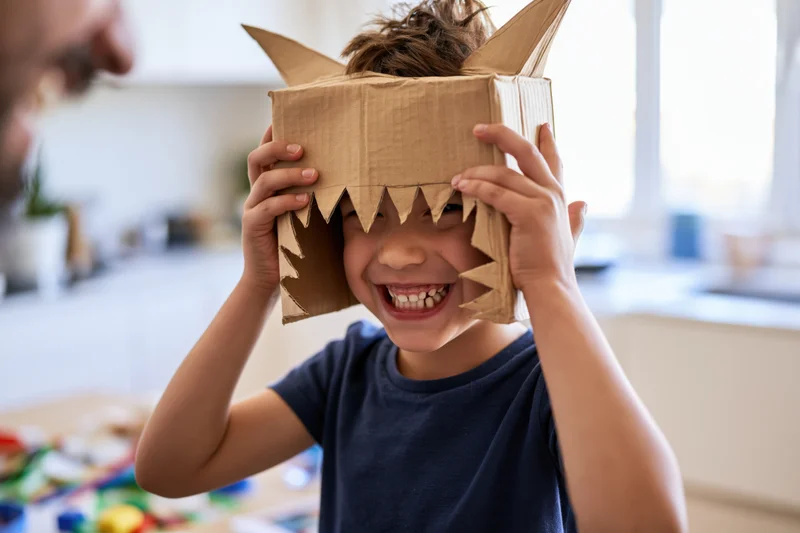 Young boy grinning through a handmade cardboard monster mask while holding it on his head in a kitchen