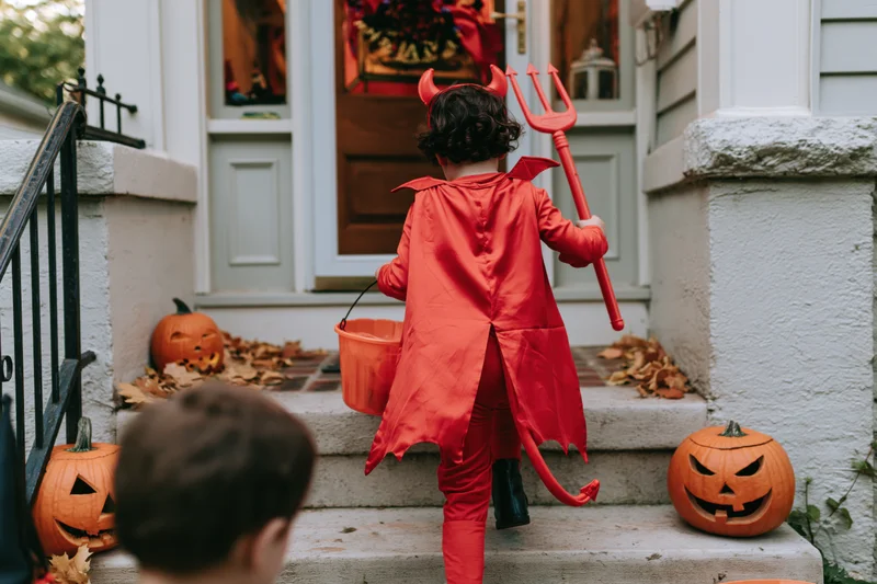 Child in red devil costume with cape and fork standing on porch steps near jack-o-lanterns for halloween trick-or-treating