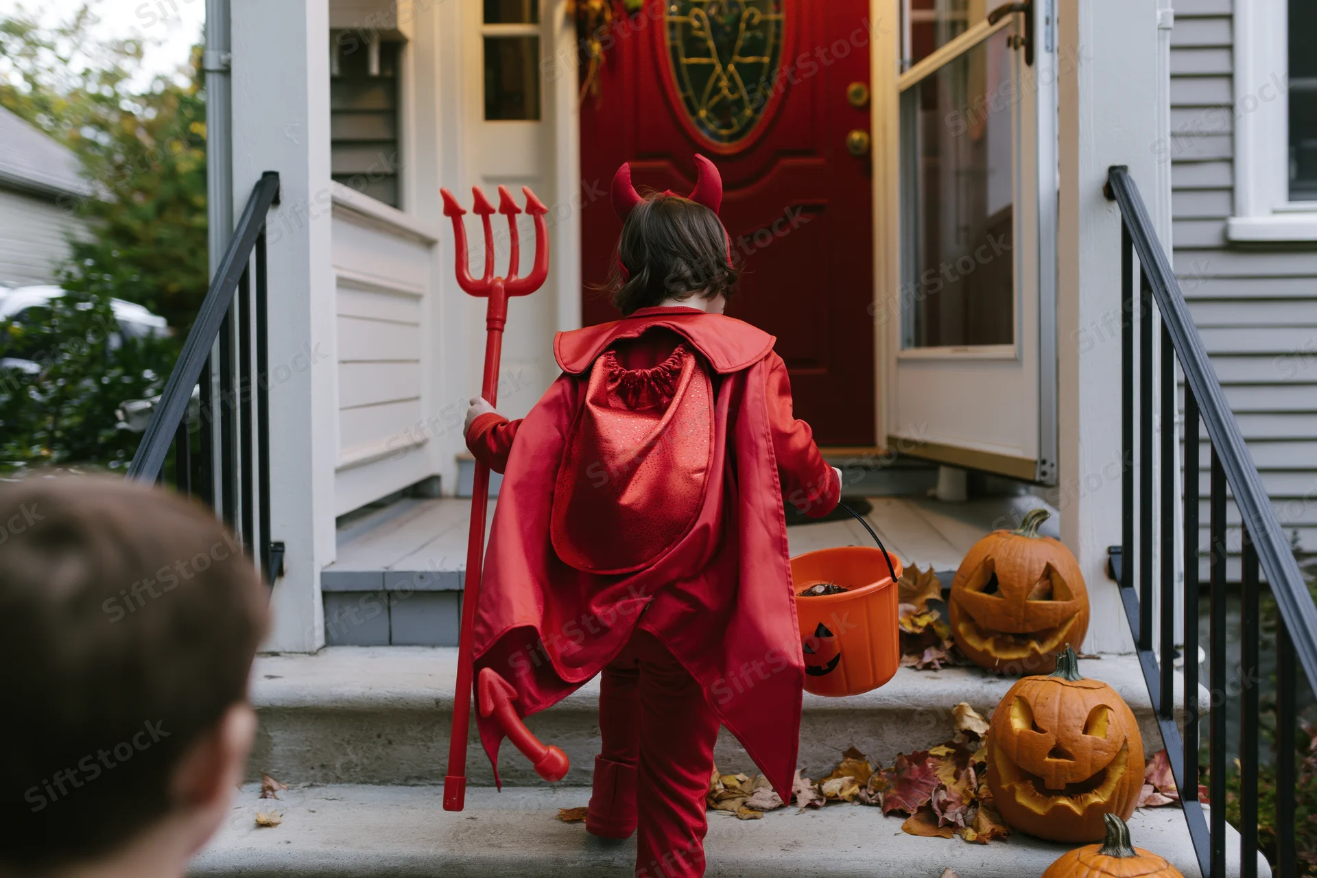 Child in red devil costume with cape and trident holding candy bucket on porch steps near carved pumpkins