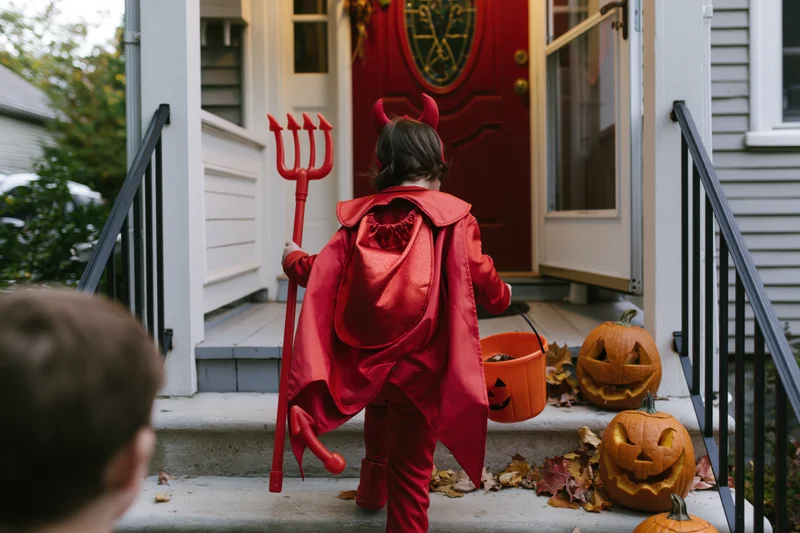 Child in red devil costume with cape and trident holding candy bucket on porch steps near carved pumpkins